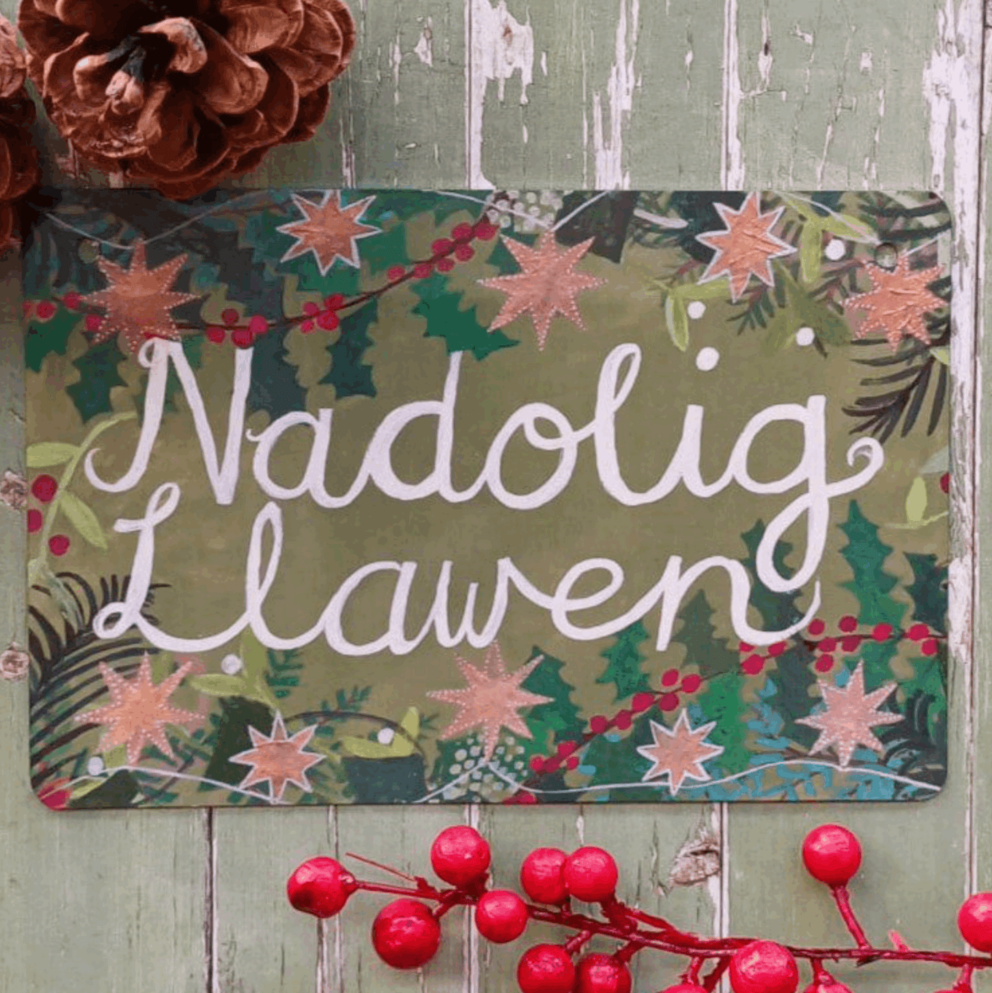 A rectangular Christmas sign with the Welsh greeting 'Nadolig Lawen' in white script over a green and pink holly pattern, displayed on a rustic green wooden surface with a pinecone and red berries as props.
