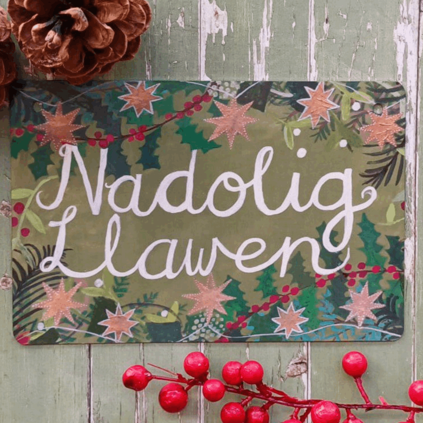 A rectangular Christmas sign with the Welsh greeting 'Nadolig Lawen' in white script over a green and pink holly pattern, displayed on a rustic green wooden surface with a pinecone and red berries as props.
